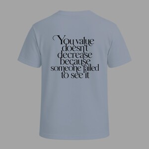 May include: A light blue t-shirt with the text "You value doesn't decrease because someone failed to see it" in black script. The shirt has short sleeves and a crew neck, suitable for casual wear.