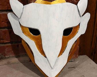 May include: A handmade mask with a white and gold color scheme. The mask has a stylized design with pointed features and large eye openings. The mask is painted with white and gold paint, and it is displayed against a brick wall.