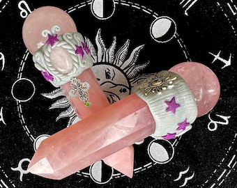 May include: Two pink rose quartz crystal wands with white and silver decorative accents. The wands are topped with a pink rose quartz crystal and have a silver charm hanging from the bottom. The wands are lying on a black background with a zodiac wheel design.