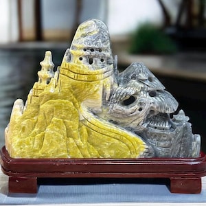 May include: A hand-carved stone sculpture of a mountain landscape with a yellow and gray color scheme. The sculpture is displayed on a wooden base.