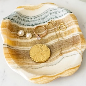 May include: A decorative onyx tray with a gold coin, a ring with a clear stone, a gold heart pendant necklace, and two pearl earrings. The tray has a natural, layered pattern of white, gold, and brown.