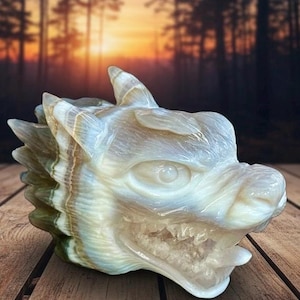 May include: A carved stone dragon head sculpture. The sculpture is primarily white with streaks of tan, green, and brown. The dragon has detailed features, including an open mouth, visible teeth, and a textured surface. The background is a blurred forest scene.