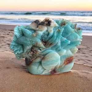 May include: A carved sculpture of aquatic life, featuring starfish, fish, and a turtle. The sculpture is primarily turquoise with brown and white accents. The artwork is set on a sandy beach with the ocean in the background.