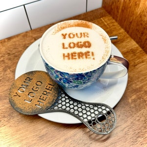 May include: A white ceramic mug with a floral pattern and a cinnamon design that says "YOUR LOGO HERE!" on top of the foam. A black metal stencil with the same text is next to the mug.