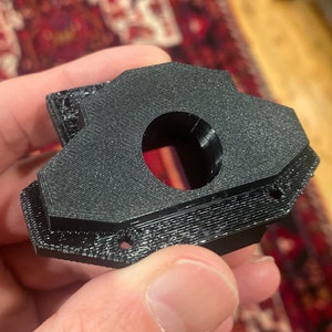 Może przedstawiać: Czarna część z tworzywa sztucznego, wydrukowana w 3D, z okrągłym otworem w środku. Część ma fakturowaną powierzchnię i wydaje się być elementem większego zespołu.