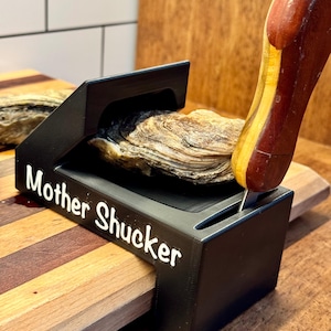 May include: A black oyster shucker tool with a wooden handle, holding an oyster on a wooden cutting board. The tool is labeled "Mother Shucker".