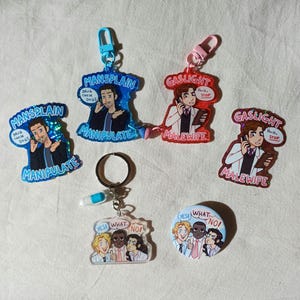 May include: A selection of acrylic keyrings and a button, each with illustrations and text. The keyrings display phrases such as "Mansplain" and "Gaslight" with cartoon characters. The button reads "Yes! What? No!" with a group of people.
