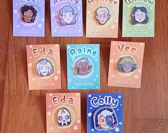 Op de afbeelding: Een verzameling karakterspelden op kaarten. Elke speld toont het gezicht van een ander personage uit de animatieserie, met namen als "Luz", "Amity" en "Hunter" erboven. De kaarten hebben een ster- en hartpatroon en de tekst "MELACHAE ART".