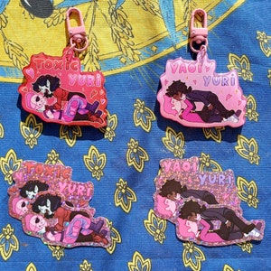 May include: Four pink and glitter keychains with cartoon couples. Two keychains feature the text "Toxic Yuri" and the other two feature the text "Yaoi Yuri".
