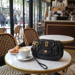 May include: A black leather handbag with gold detailing, a coffee with latte art, and a croissant on a marble table in a cafe. Wicker chairs and a street scene are visible in the background. The table is approximately 60 cm in diameter.