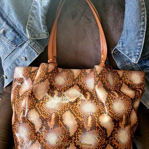 May include: A brown and tan snakeskin-patterned tote bag with brown leather handles. The bag has the brand name "Dooney & Bourke" printed on the front. A blue denim jacket is behind the bag.