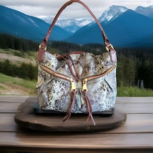 May include: A snakeskin-patterned handbag with a brown leather strap and gold zippers. The bag has a drawstring closure and is set against a backdrop of mountains and a wooden surface.