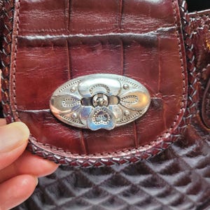 May include: A close-up of a burgundy leather purse with a silver clasp. The purse has a quilted pattern and a braided trim.