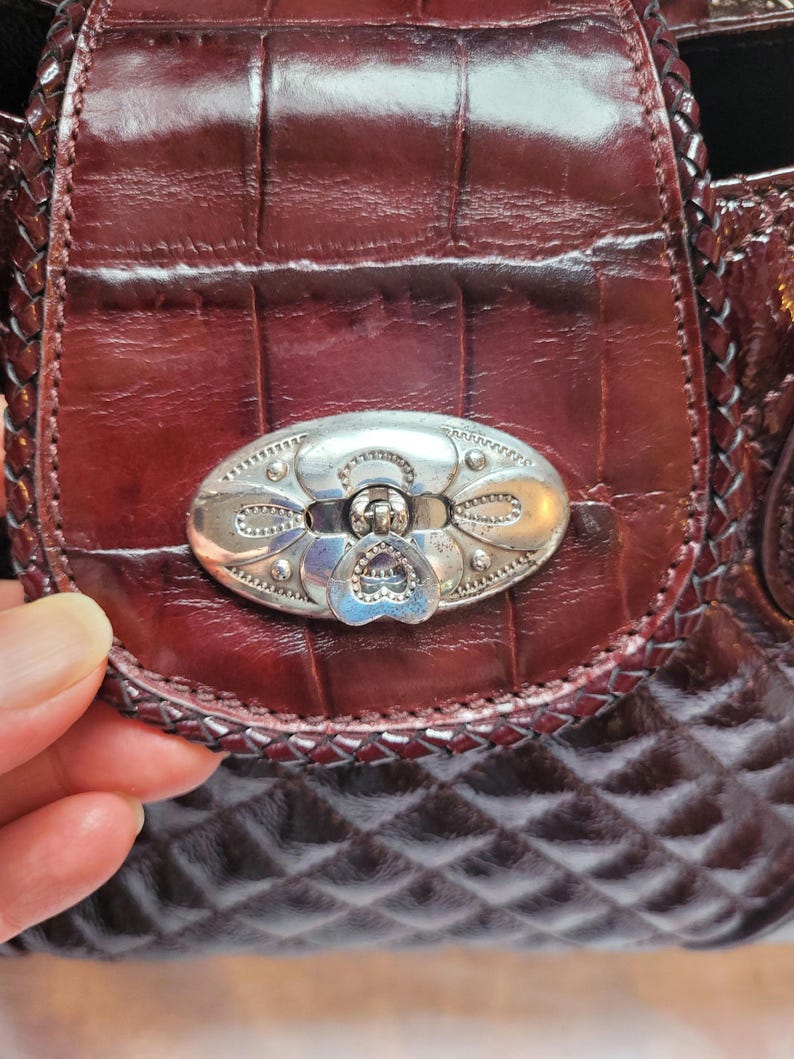 May include: A close-up of a burgundy leather purse with a silver clasp. The purse has a quilted pattern and a braided trim.