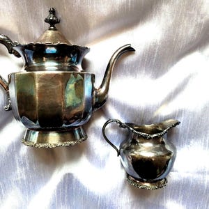 Könnte beinhalten: Ein silbernes Teeservice, bestehend aus einer Teekanne mit Deckel und einem Sahnekännchen. Die Teekanne hat einen gebogenen Griff und Ausguss, und das Sahnekännchen hat einen kleinen Griff. Beide Teile haben verzierte Details am Boden und am Rand.
