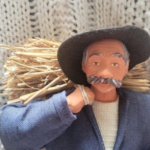 May include: A figurine of an elderly man with a gray mustache and a dark hat, carrying a bundle of straw on his shoulder. He wears a blue jacket over a white sweater. The figurine is set against a textured, off-white background.