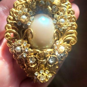 Puede incluir: Broche ovalado dorado con una gran perla central. El broche presenta intrincados diseños florales y de volutas, acentuados con pequeñas perlas y cristales transparentes. Una joya de inspiración vintage.