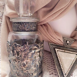 May include: A clear glass jar with a decorative silver metal base and lid. The jar has a clear glass stopper. A silver and white triangle-shaped brooch is next to the jar.