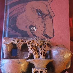 Puede incluir: Un libro con la palabra "GRYFFINDOR" en letras doradas, con una ilustración de un león. En primer plano, se ve una escultura de piedra tallada con elementos florales y arquitectónicos. La paleta de colores incluye tonos de rojo, marrón y dorado.