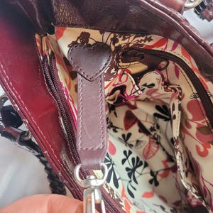 May include: A burgundy leather handbag with a floral patterned lining. The bag has a zipper closure and a heart-shaped leather pull tab.