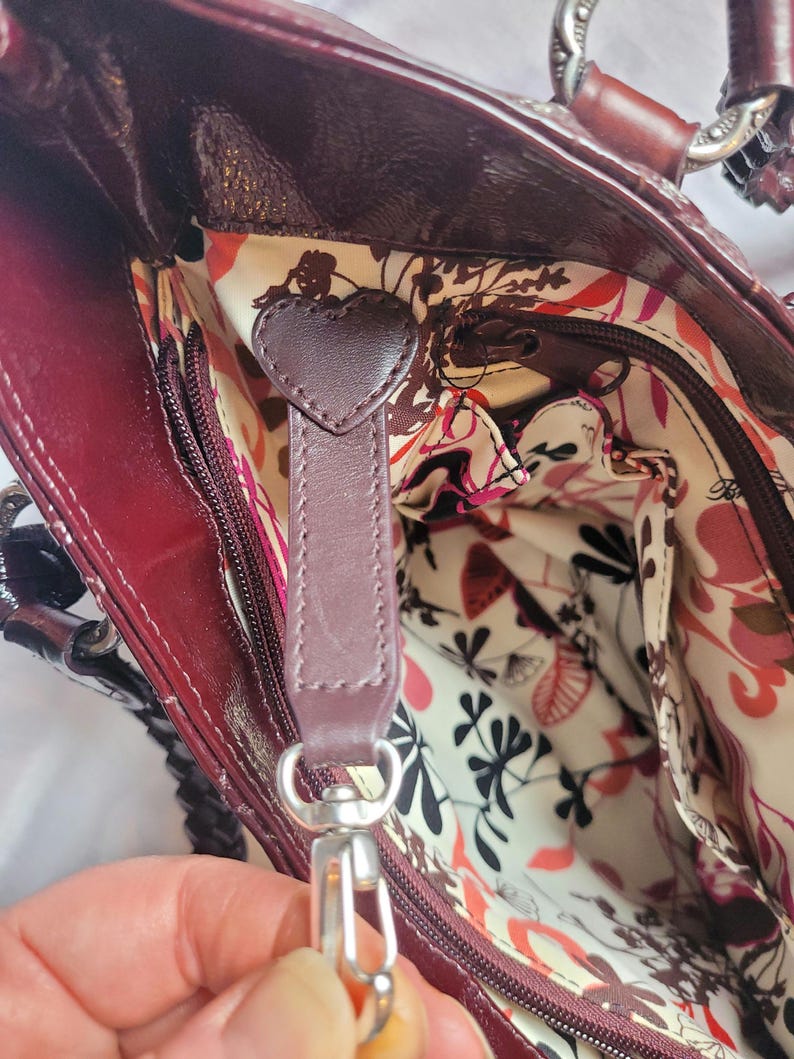 May include: A burgundy leather handbag with a floral patterned lining. The bag has a zipper closure and a heart-shaped leather pull tab.
