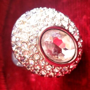 Puede incluir: Un anillo plateado con una gran gema ovalada y transparente en el centro, rodeada de piedras claras más pequeñas y brillantes. El anillo se encuentra sobre un fondo rojo intenso, creando un contraste llamativo.