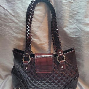 May include: A brown leather handbag with a quilted design and a braided strap. The handbag has a flap closure with a decorative buckle.
