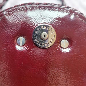 May include: Close-up of a burgundy leather purse with a silver metal clasp that reads "Brighton" in a circle.