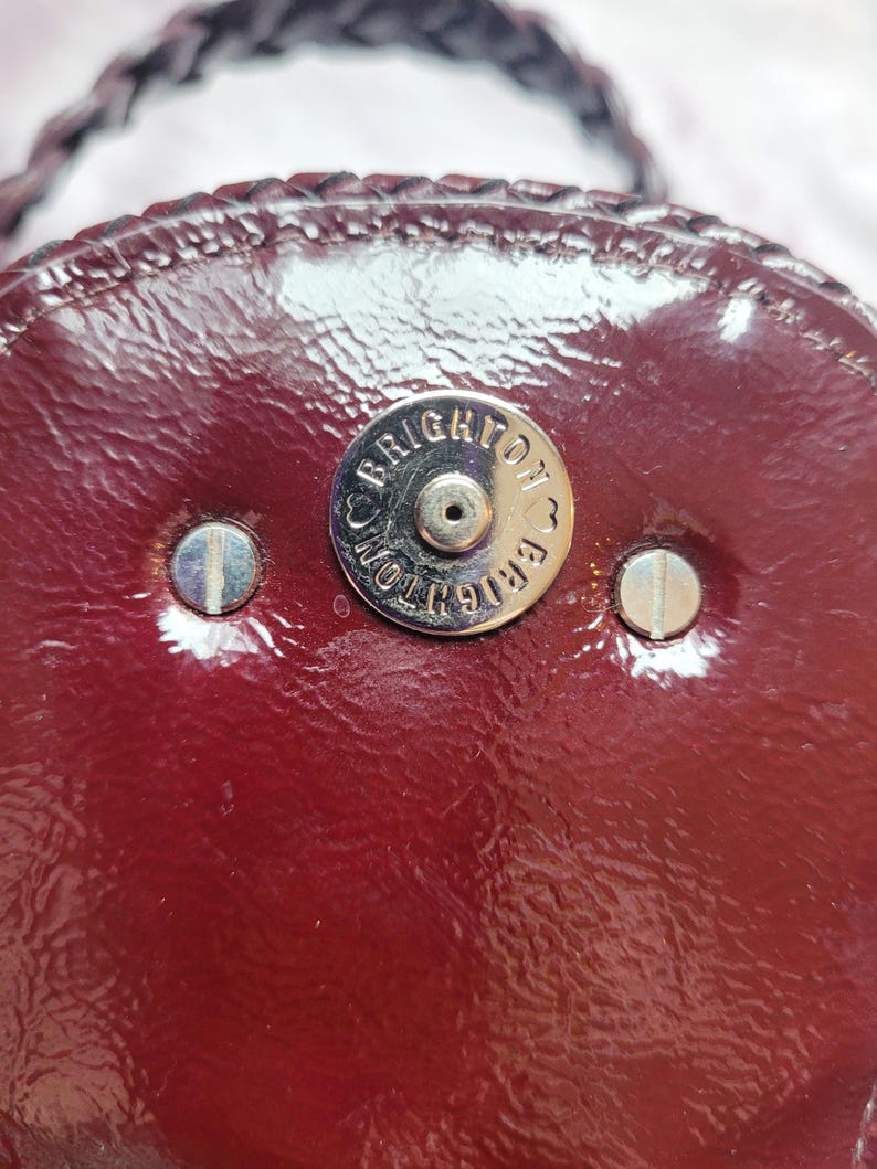May include: Close-up of a burgundy leather purse with a silver metal clasp that reads "Brighton" in a circle.