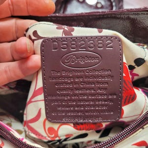 May include: A close-up of a burgundy leather handbag with a floral lining. The interior tag reads "D582382 Brighton The Brighton Collection handbags are individually crafted in China from quality leathers. Any markings on the surface are part of the natural beauty, texture and character of the leather, which make each bag unique."