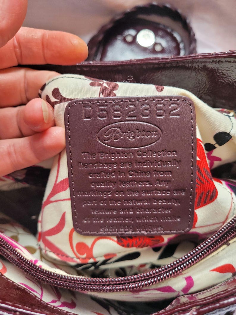 May include: A close-up of a burgundy leather handbag with a floral lining. The interior tag reads "D582382 Brighton The Brighton Collection handbags are individually crafted in China from quality leathers. Any markings on the surface are part of the natural beauty, texture and character of the leather, which make each bag unique."