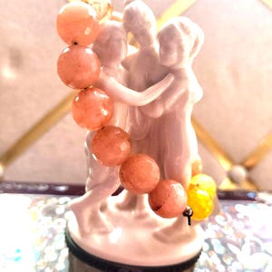 May include: A white ceramic figurine of three embracing figures, with a strand of peach-colored beads draped over them. The base of the figurine is a small box with the text "Hearst Castle".