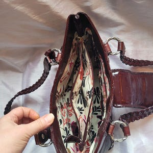 May include: A brown leather handbag with a braided strap and a floral patterned lining. The bag has multiple compartments and a zipper closure.