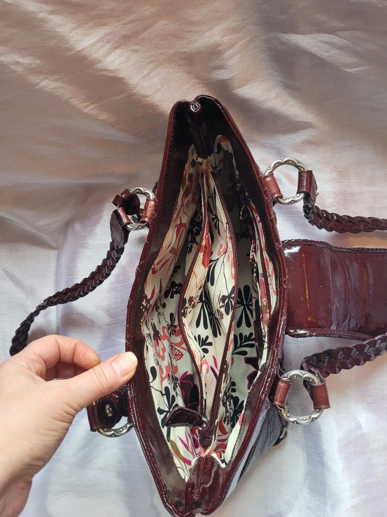 May include: A brown leather handbag with a braided strap and a floral patterned lining. The bag has multiple compartments and a zipper closure.