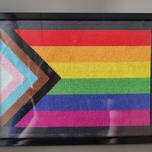 May include: Framed cross-stitch artwork featuring the Progress Pride flag. The flag design includes the traditional rainbow stripes, along with a chevron of colors representing transgender, marginalized, and people of color. The artwork is encased in a black frame.