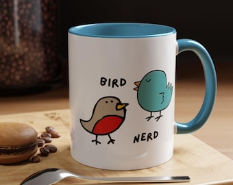 May include: White ceramic mug with a blue rim and handle. The mug features a cartoon bird with red breast and the word "bird" above it. A second cartoon bird in blue is next to it with the word "nerd" below it.