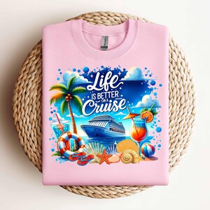以下が含まれることがあります： クルーズ船、ヤシの木、カクテル、そして「Life is better on a cruise」というテキストが描かれたカラフルなグラフィックデザインのピンクのTシャツです。