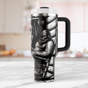 May include: A stainless steel travel mug with a black and white graphic of a Bigfoot sitting in a rocking chair. The mug has a handle and a lid.