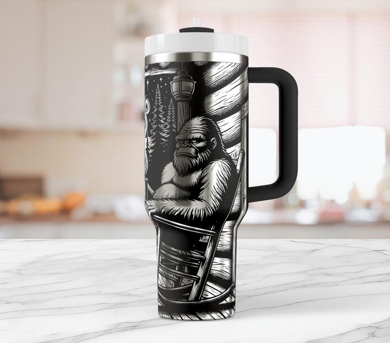 May include: A stainless steel travel mug with a black and white graphic of a Bigfoot sitting in a rocking chair. The mug has a handle and a lid.