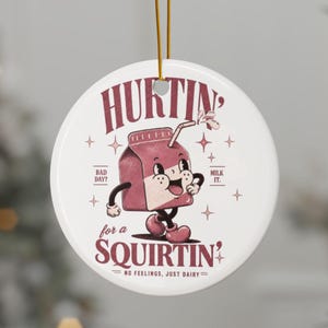May include: A round white ceramic ornament featuring a cartoon pink milk carton character with arms and legs. The text reads HURTIN' for a SQUIRTIN', BAD DAY? MILK IT. NO FEELINGS, JUST DAIRY. It hangs from a gold string against a blurred background.