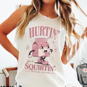 May include: An ivory sleeveless tank top featuring a graphic of a pink milk carton character with arms and legs. The text reads HURTIN' for a SQUIRTIN', BAD DAY?, MILK IT., and NO FEELINGS, JUST DAIRY. The design includes small star accents.