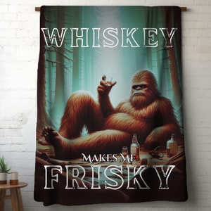 May include: A brown and orange Bigfoot character sitting in a forest setting with a glass of whiskey in one hand and a bottle of whiskey in the other. The text "Whiskey Makes Me Frisky" is printed on the image.