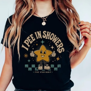 May include: A black t-shirt featuring a graphic of a yellow cartoon star character with arms and legs, giving a thumbs up. The text reads I PEE IN SHOWERS and FIVE STAR HABIT, surrounded by small star icons and a checkered pattern at the base.