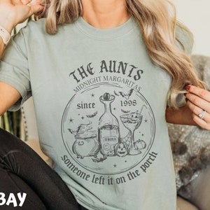 K&ouml;nnte beinhalten: Ein hellblaues T-Shirt mit einem schwarz-wei&szlig;en Grafikdesign. Die Grafik zeigt einen Kessel, eine Flasche Tequila, ein Margarita-Glas, Flederm&auml;use und Sterne. Der Text auf der Grafik lautet "The Aunties Midnight Margaritas since 1998 someone left it on the porch".