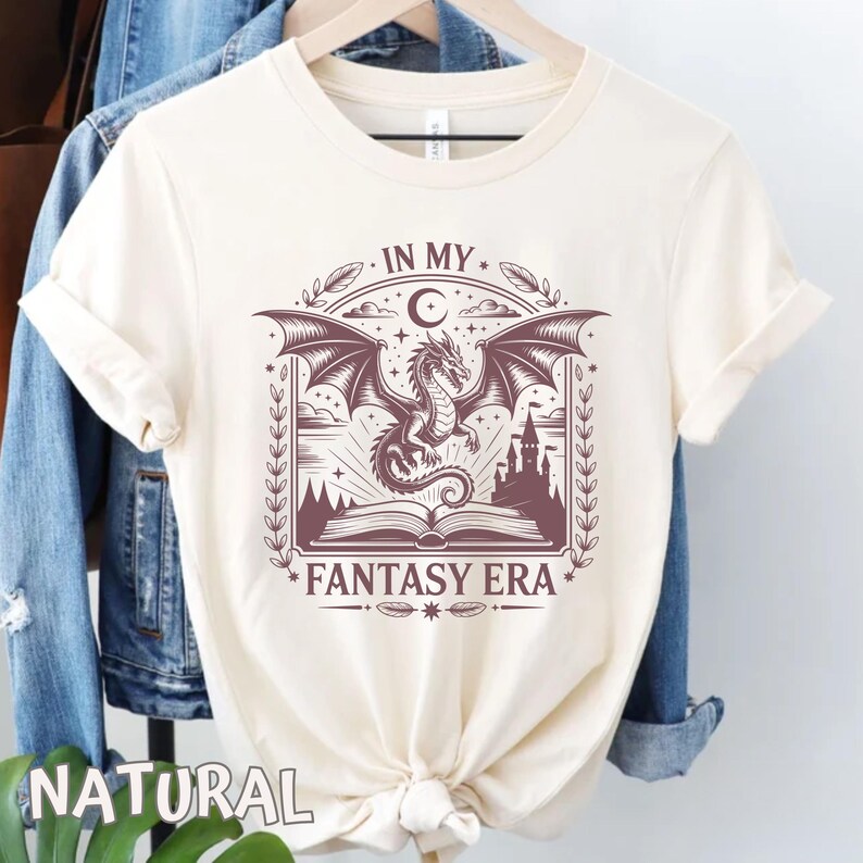 May include: A cream-colored short-sleeve t-shirt featuring a dark brown graphic print of a dragon flying above an open book and a castle. The text "IN MY FANTASY ERA" is printed in a decorative font above and below the central illustration.
