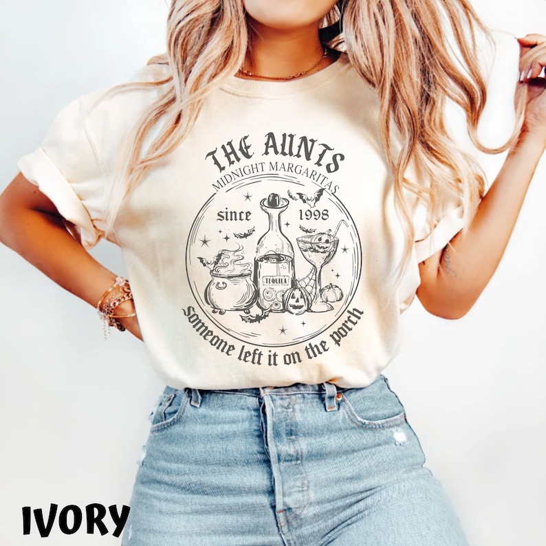 K&ouml;nnte beinhalten: Ein T-Shirt in Elfenbein mit einem schwarz-wei&szlig;en Grafikdesign. Die Grafik zeigt einen Kessel, eine Flasche Tequila, ein Cocktailglas und Flederm&auml;use. Der Text lautet "The Aunties Midnight Margaritas since 1998 Someone left it on the porch."