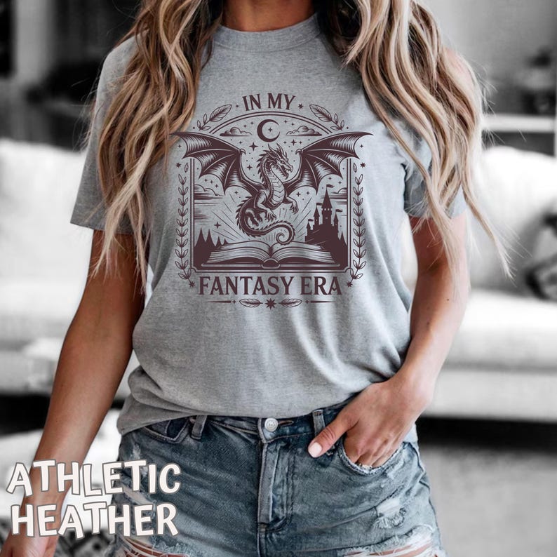May include: A heather gray short-sleeve t-shirt featuring a dark brown graphic print of a dragon flying above an open book and a castle. The text "IN MY FANTASY ERA" is printed in a decorative font around the central illustration.