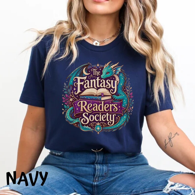 May include: A navy blue short-sleeve t-shirt featuring a graphic print of a teal dragon curled around an open book. The text reads The Fantasy Readers Society in a decorative font, surrounded by colorful floral elements, stars, and a crescent moon.