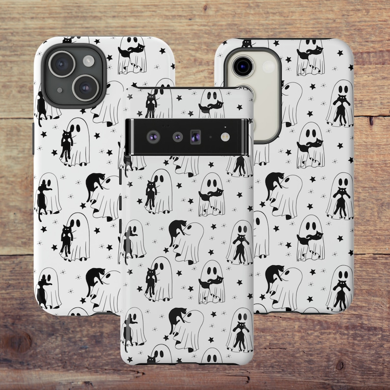 May include: Two white phone cases with a black and white pattern of ghosts and cats. The cases have a white background with black stars and black outlines of ghosts and cats. The ghosts are holding cats in their arms. The cases are for a phone with a triple camera lens.