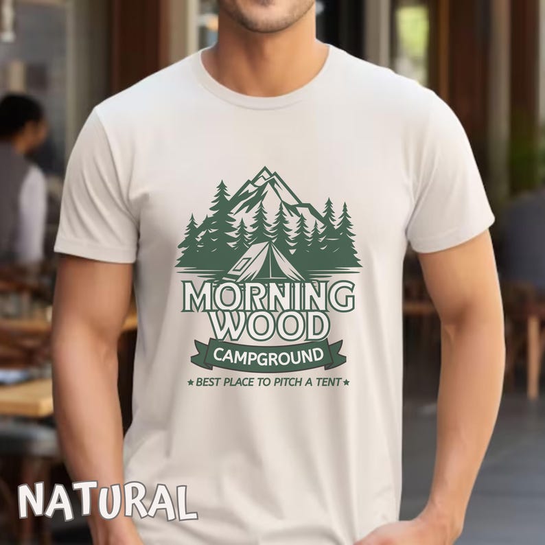 Puede incluir: Una camiseta blanca con un dise&ntilde;o gr&aacute;fico verde de una cordillera, &aacute;rboles y una tienda de campa&ntilde;a. El texto "Morning Wood Campground" est&aacute; impreso en verde debajo del gr&aacute;fico. El texto "Best Place to Pitch a Tent" est&aacute; impreso en verde m&aacute;s peque&ntilde;o debajo del nombre del camping.